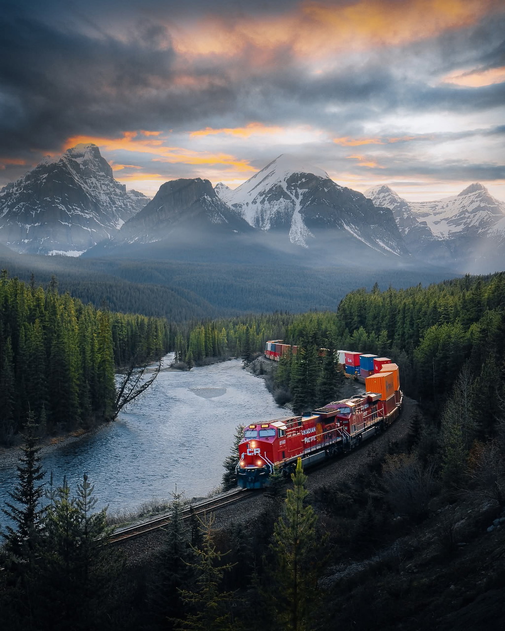 Freight train curving along a river through pine forest with snow-capped mountains and dramatic sunset clouds