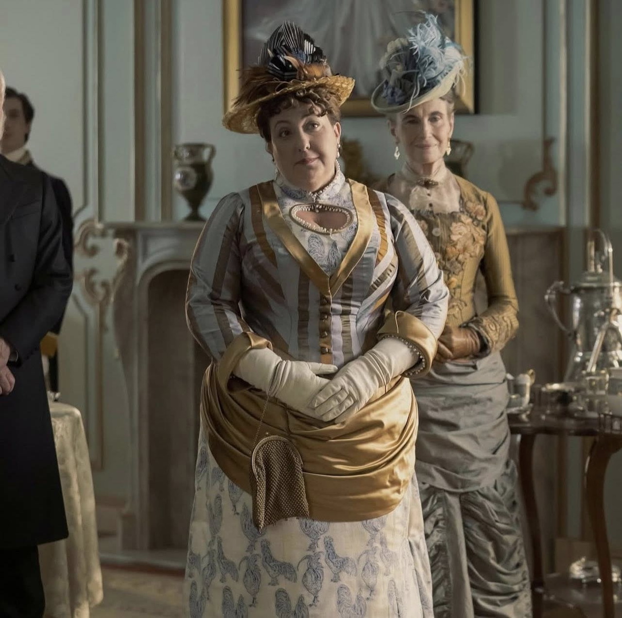 Woman in an Edwardian gown and hat standing in a vintage interior with another woman behind her