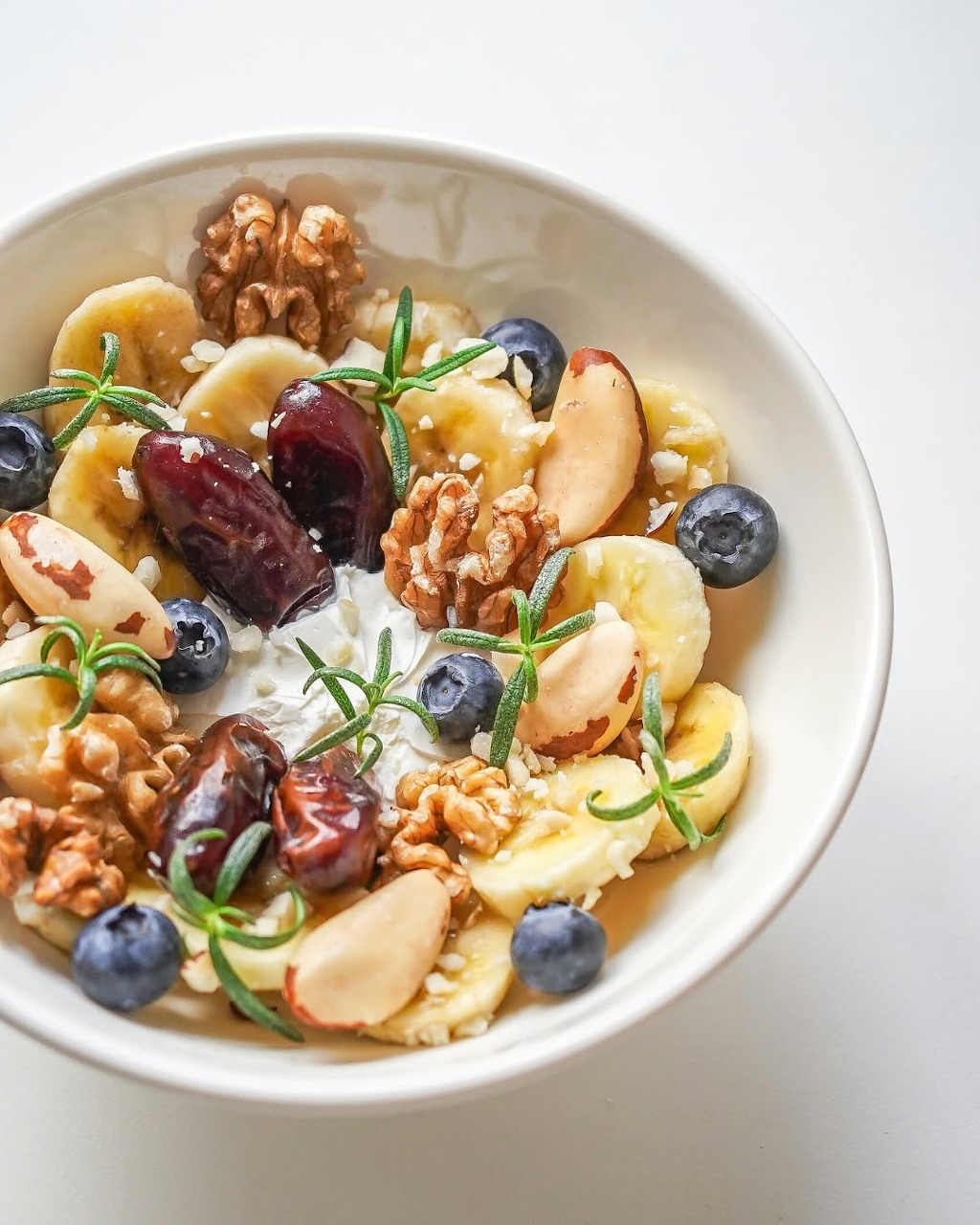 White bowl with banana, blueberries, cherries, apple, walnuts, yogurt and rosemary