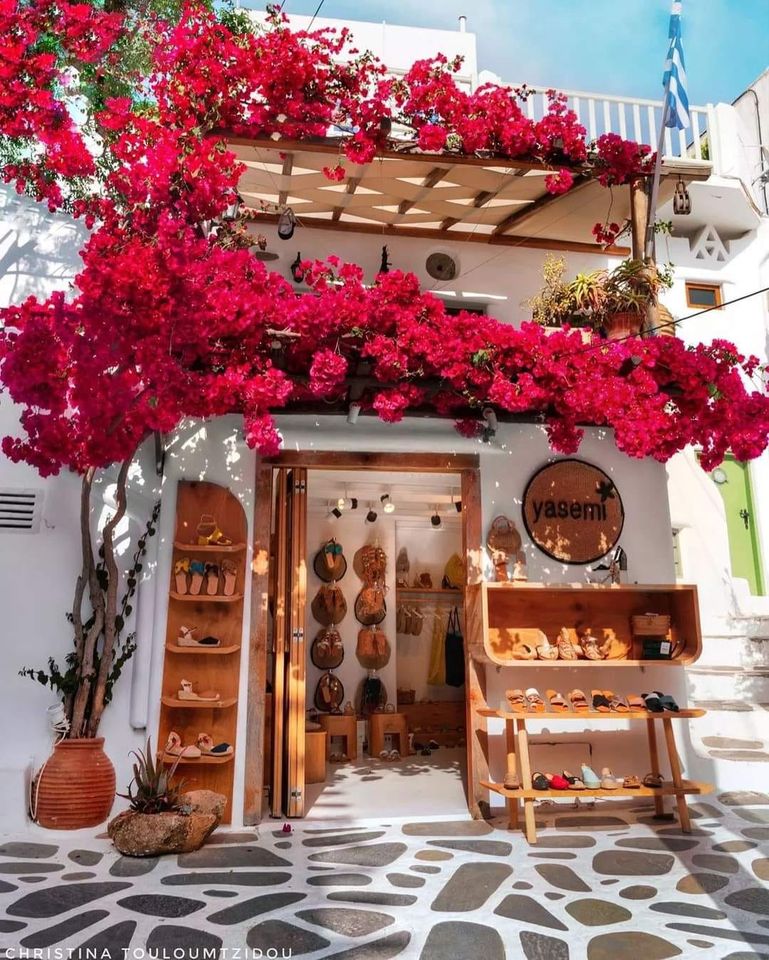 White Greek shop entrance covered in bright pink bougainvillea flowers with handcrafted items displayed outside