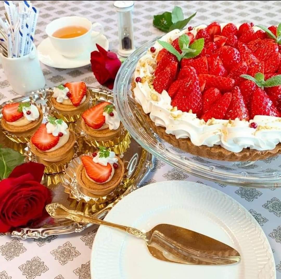 Strawberry cream cake on glass stand with mini fruit tartlets, roses, and tea cups on elegant table setting