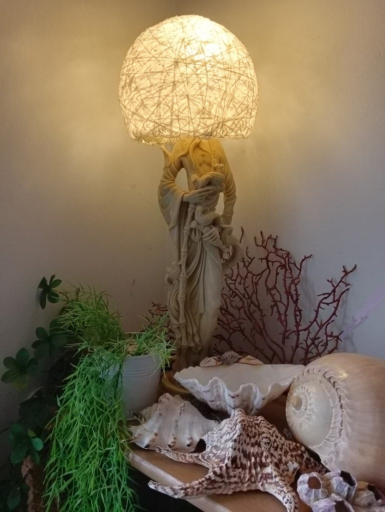 Decorative lamp with dome shade and statue base surrounded by shells, coral and plants on wooden table