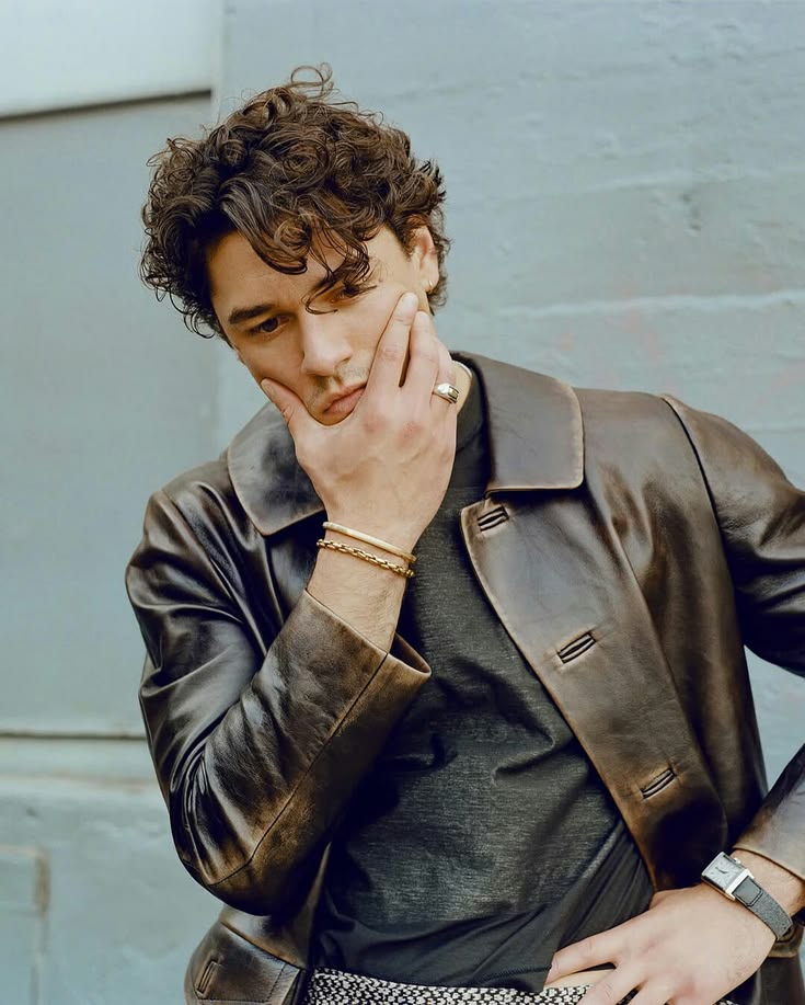 Young man with curly hair in a brown leather jacket posing with hand near chin, wearing gold bracelets