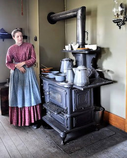 Woman in 19th century dress next to ornate black cast-iron stove in vintage kitchen