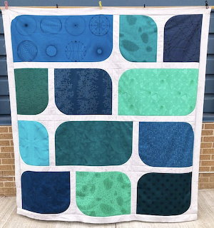 Patchwork quilt with blue, teal, and green rounded rectangular blocks on white background hung on a fence