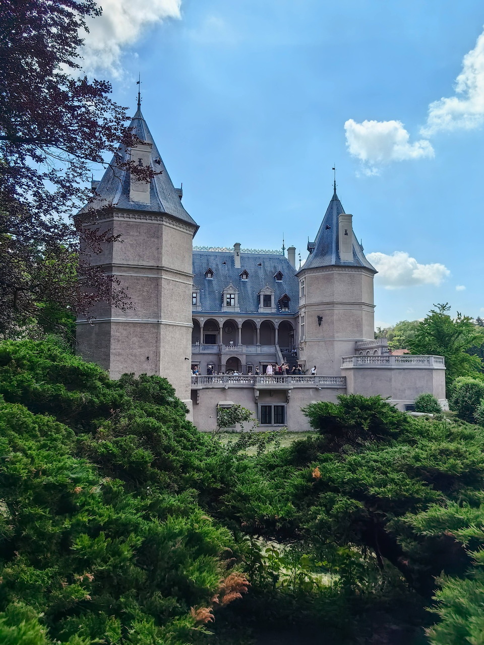 Medieval castle with stone towers and mansard roof surrounded by green trees under a blue sky
