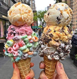 Two hands holding waffle cones with stacked ice cream scoops and candies against a city backdrop