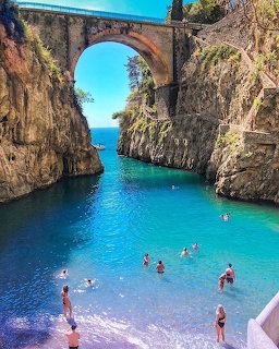 Swimmers in a turquoise rocky cove beneath a stone arch bridge on the Amalfi Coast
