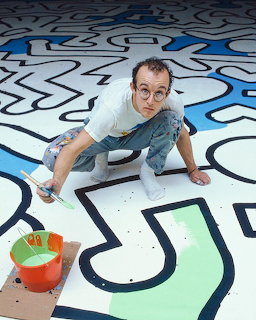 Keith Haring crouching on a colorful floor mural painting bold outlined figures with a brush