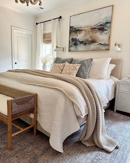 Cozy bedroom with neutral beding, layered blankets, decorative pillows, abstract painting, and woven bench