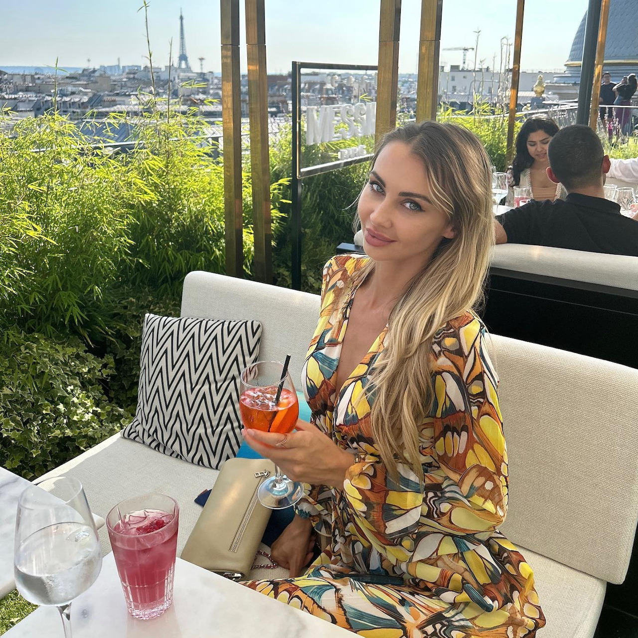 Blonde woman in floral dress smiling at outdoor café table with cocktail and city view
