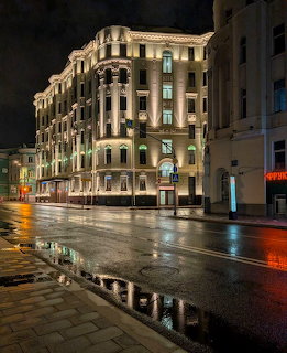 Illuminated ornate corner building at night with wet street reflections and dark stormy sky