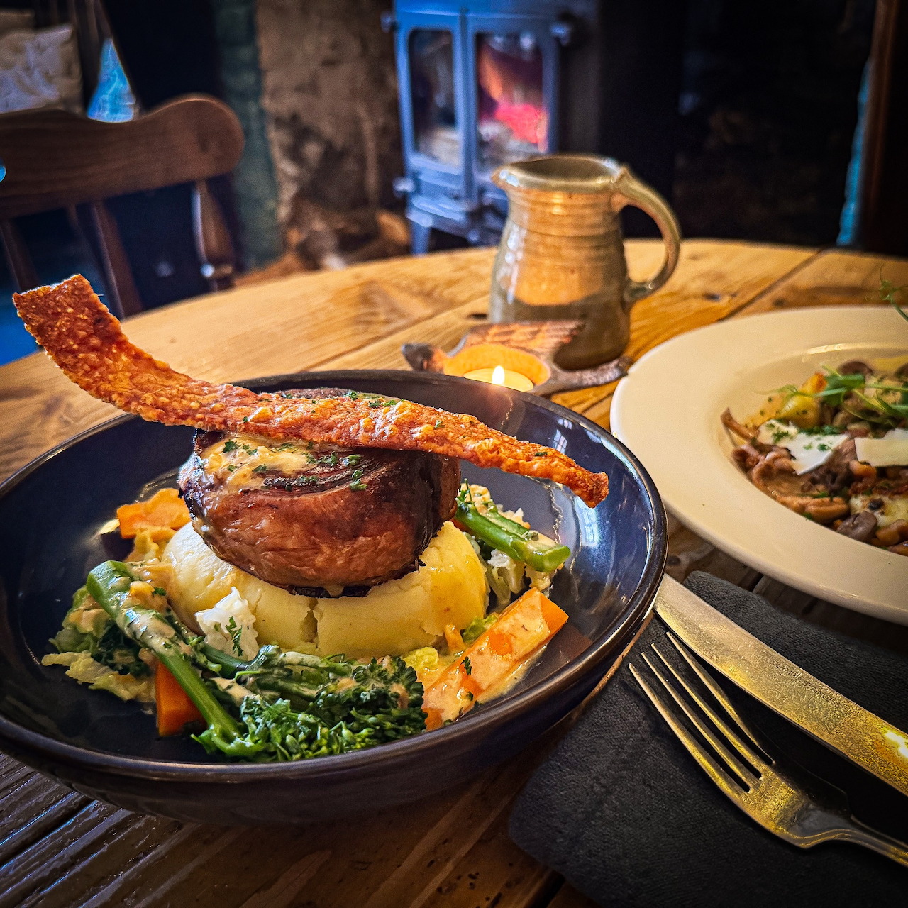 Plated pork chop with bacon strip on vegetables in black bowl, salad and mug on rustic wooden table