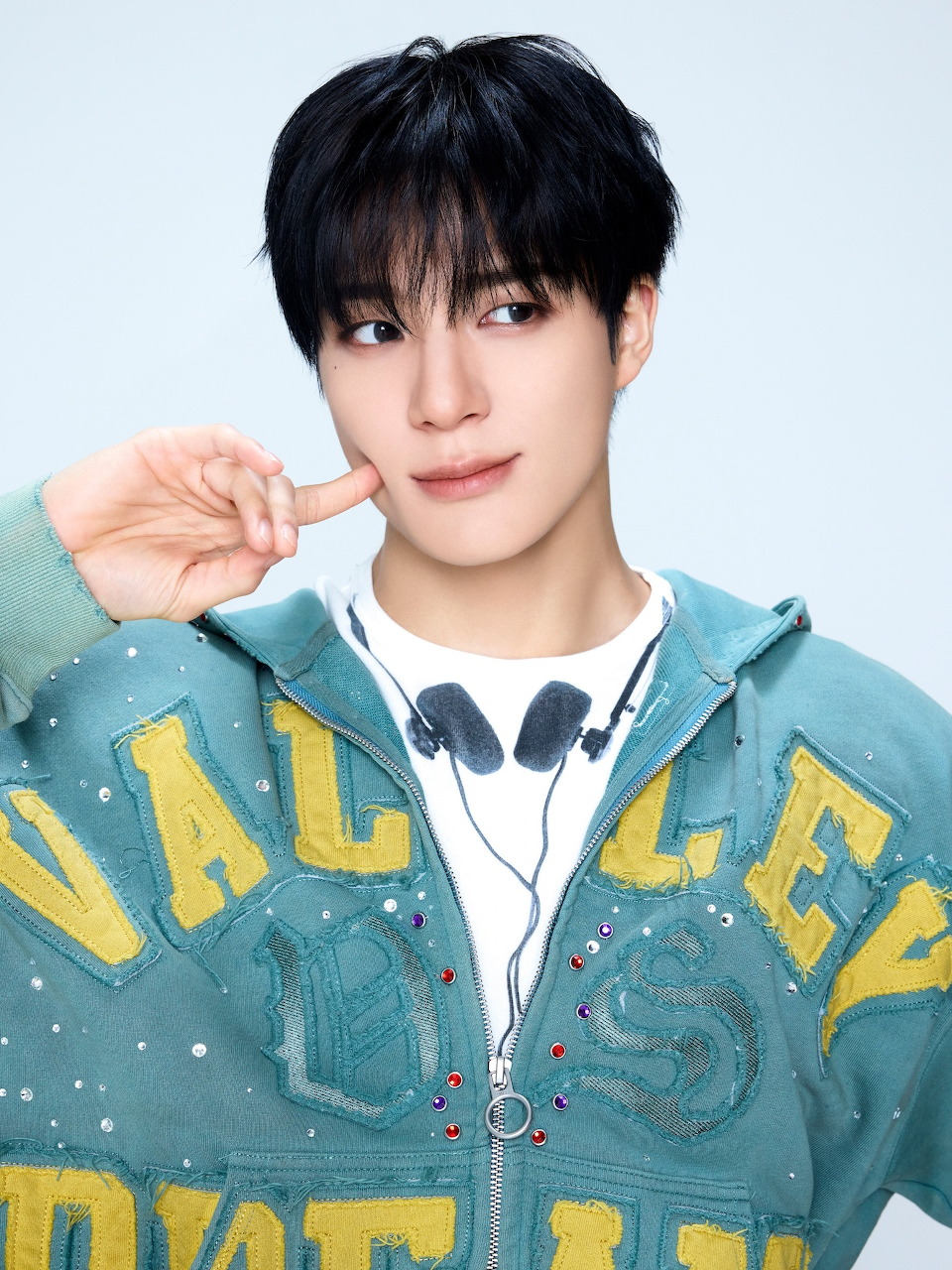 Young Korean male with black hair in a teal hoodie and headphones, making a hand gesture near his face