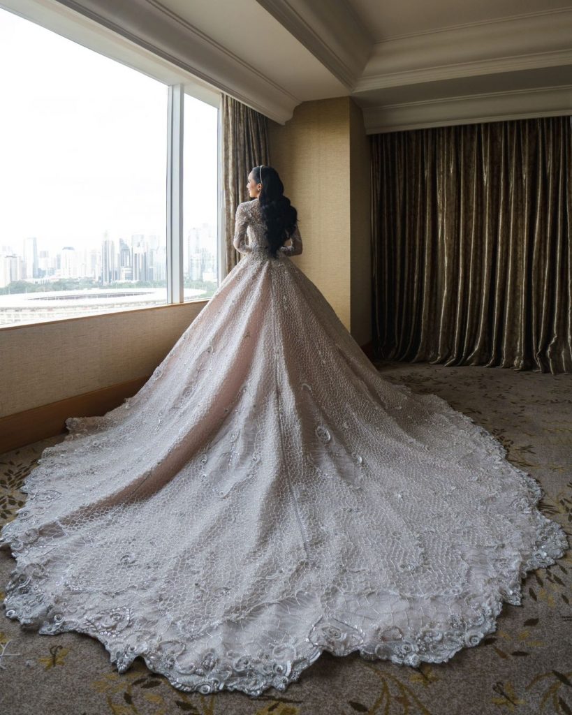Bride in elaborate white lace wedding dress with cathedral train by window with city view