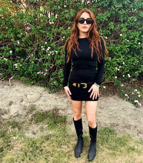 Woman in black outfit and sunglasses standing outdoors near flowering bushes