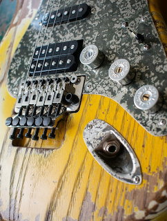 Close-up of a distressed electric guitar body with bridge, pickups, knobs, and worn yellow paint.