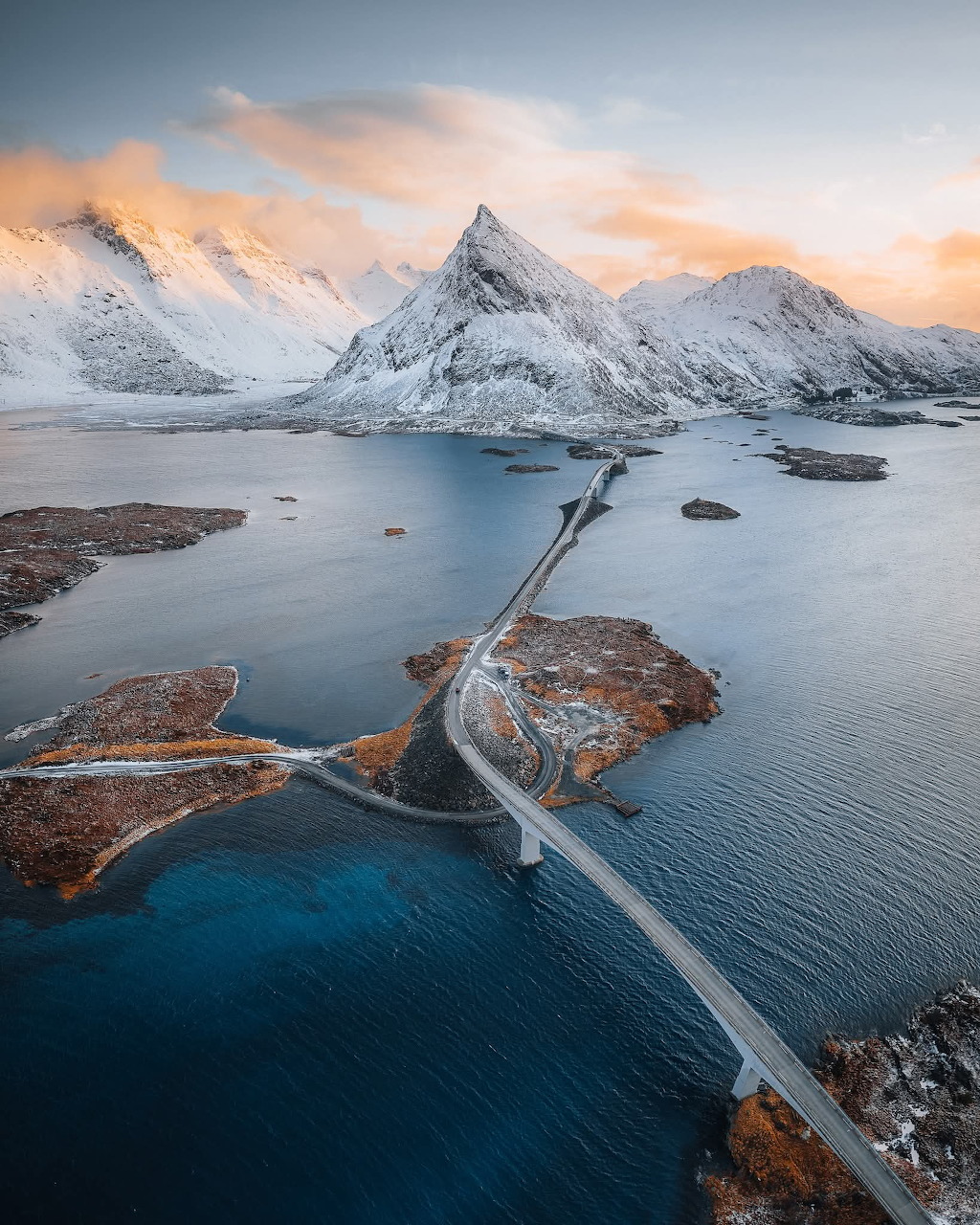 Aerial view of a bridge over a Norwegian fjord with snow-covered mountains at sunset