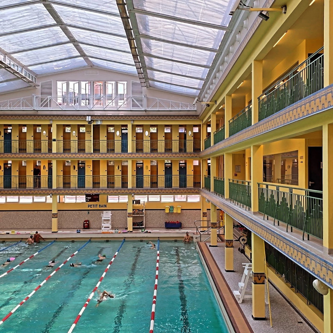 Art Deco indoor pool with glass roof, yellow walls, green railings, and swimmers in turquoise water