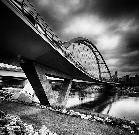 Black and white photo of a modern arch bridge over a river with dramatic clouds