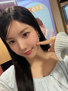 Young Korean woman taking a selfie with a peace sign gesture, wearing a light grey knit top