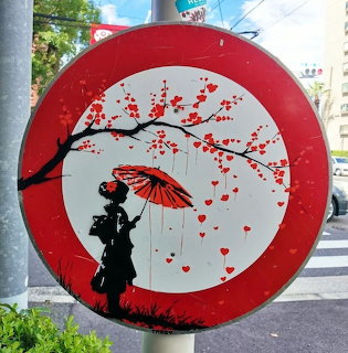 Round street art sign with a silhouette girl holding a red umbrella under a branch with red hearts