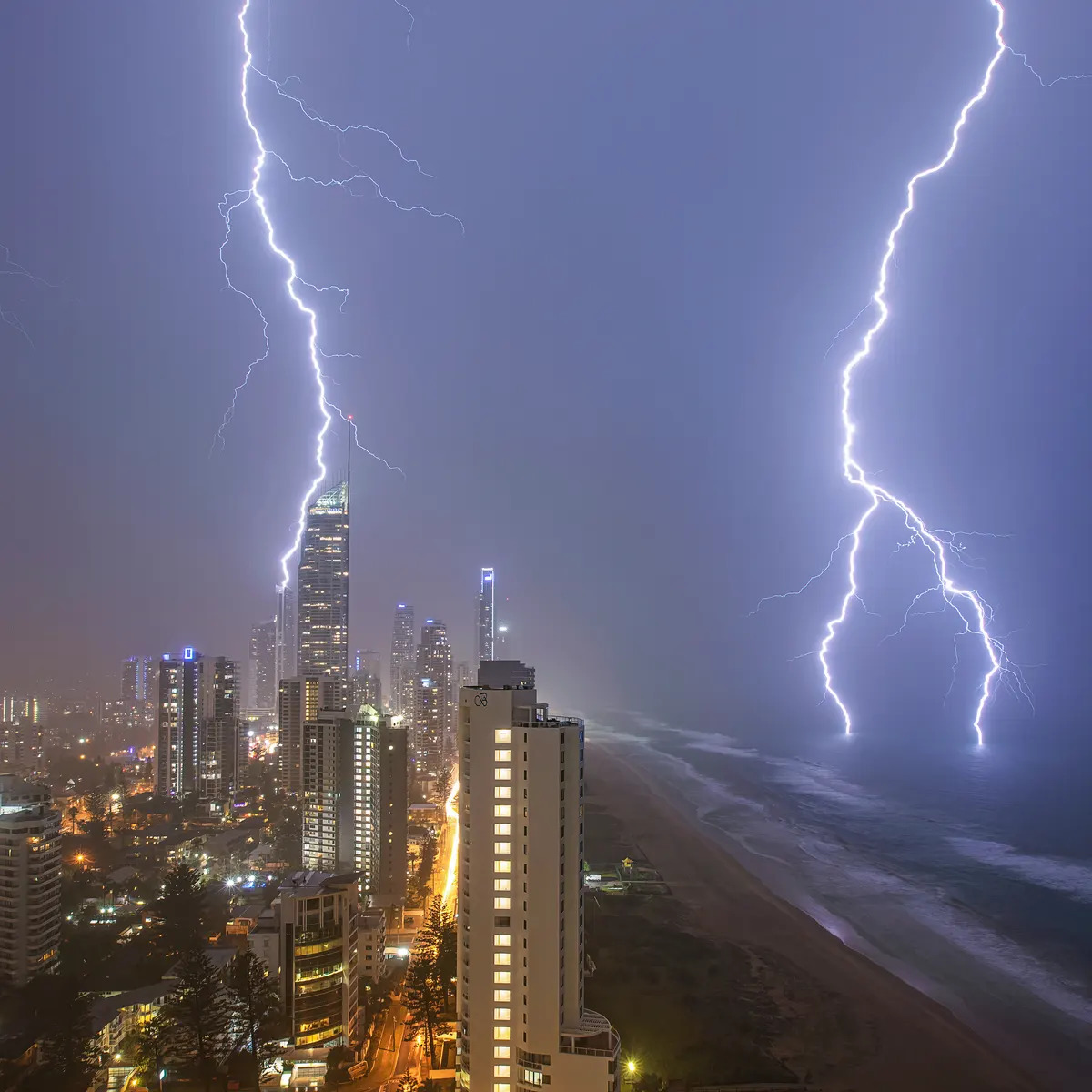 Two lightning bolts striking near lit skyscrapers along a beach at night under purple storm clouds