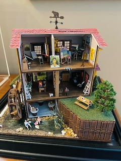 Detailed miniature dollhouse with multiple furnished rooms, tiny figures, garden area and pink roof