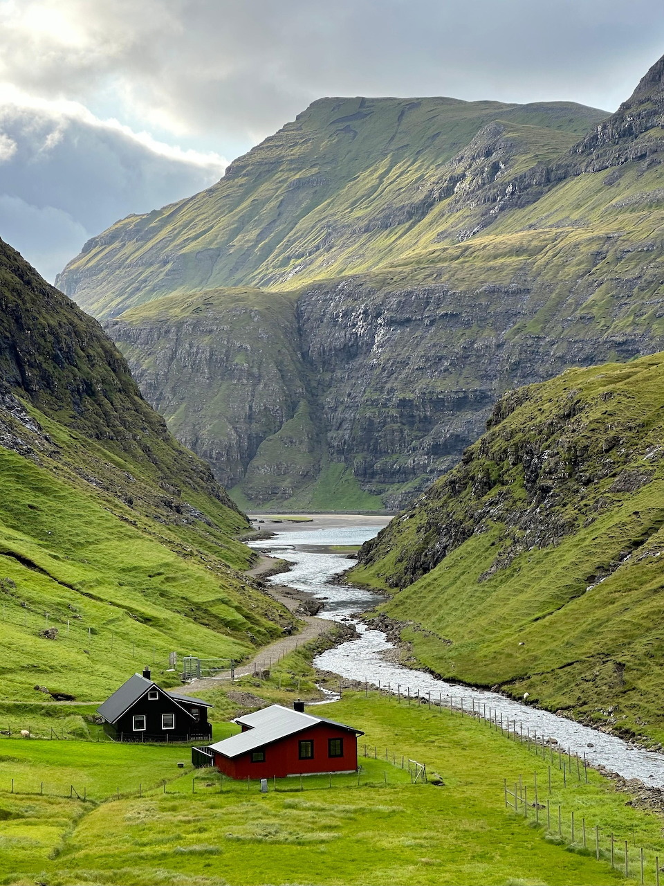 Two wooden cabins in a lush green mountain valley with a stream and lake, surrounded by steep slopes