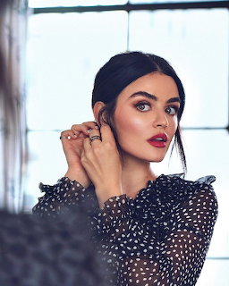 Lucy Hale wearing a black polka-dot blouse with red lipstick and hands raised near her ear