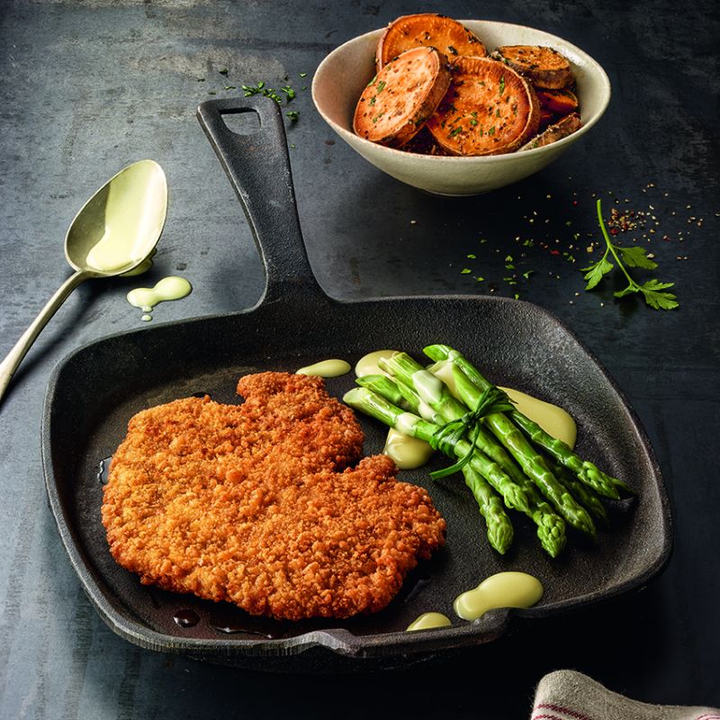 Crispy breaded schnitzel in a cast iron pan with asparagus and sweet potato chips on a dark surface