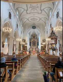 Ornate Baroque church interior with vaulted white ceilings, chandeliers, gilded altar, and wooden pews