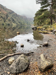 Mountain lake with rocky shore, trees, and misty hills reflected in calm water