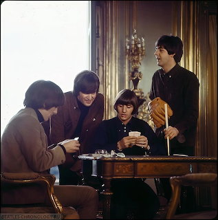 Four young men in 1960s clothing seated and standing around a table in an ornate room