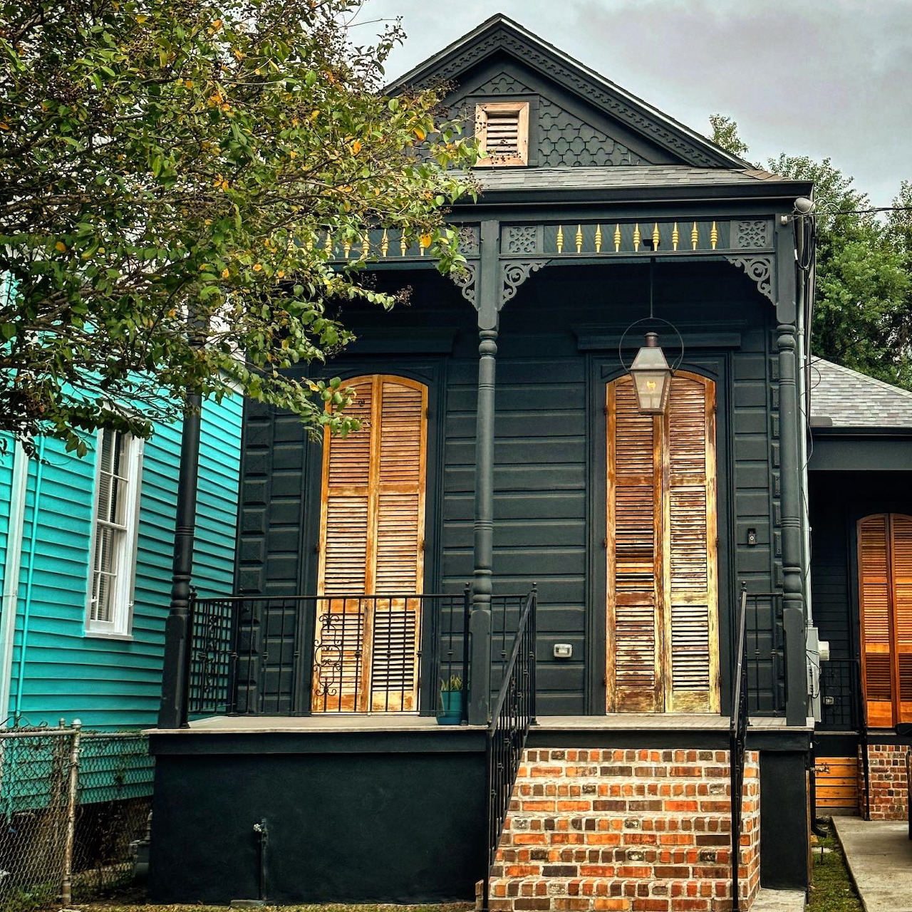 Dark gray Victorian shotgun house with yellow wooden shutters and a decorative porch on a residential street