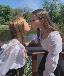 Two women in white tops kissing outdoors by a lake with trees in background