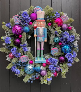 Colorful Christmas wreath with nutcracker figurine center, purple ornaments, blue flowers, and evergreen on wood wall