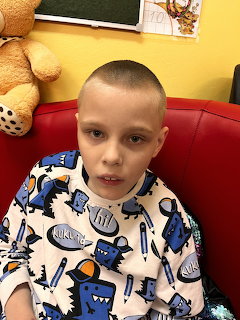 Young boy with buzz cut sitting in red barber chair wearing white cape with blue robot print