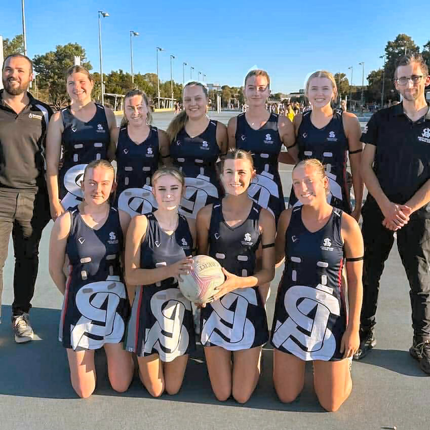 Netball team and coaches posing on an outdoor court in matching dark uniforms