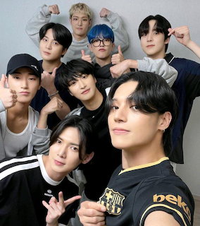 Eight young men from K-pop group Ateez posing together for a group selfie with hand gestures