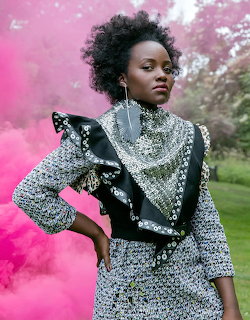 Lupita Nyong'o posing outdoors in a patterned outfit with bright pink smoke behind her
