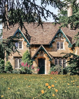 Stone cottage with steep roof and ornate gables surrounded by lush garden and tree branches
