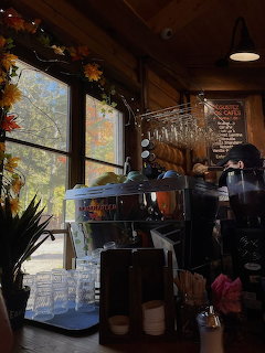 Inside a cozy wooden coffee shop with espresso machine, pastries, and fall decorations by large windows