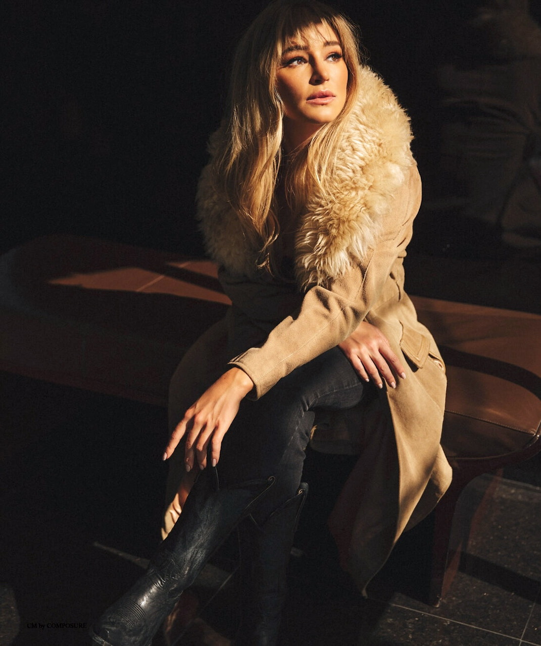 Woman with blonde bangs sitting in a tan fur-collared coat, dark jeans, and black boots in dim lighting