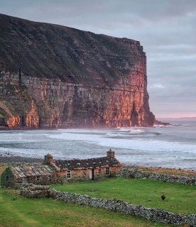 Stone cottage below towering red cliffs at Rackwick Bay with ocean waves and green grass in foreground
