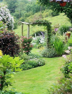 Garden path through wooden archway covered in roses, bordered by colorful flower beds and green lawn