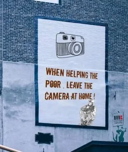 Banksy street art mural on brick wall with camera drawing and text about helping the poor