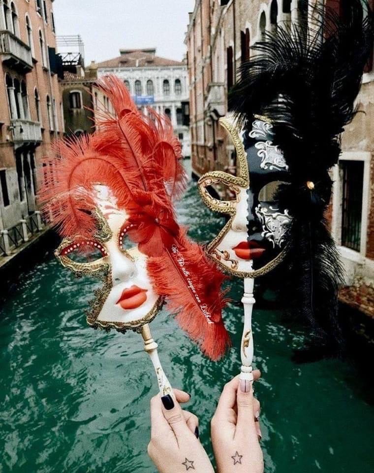 Two elaborate Venetian carnival masks with red and black feathers held over a Venice canal