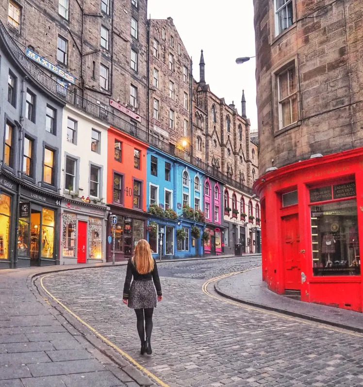 Person walking on cobblestone Victoria Street in Edinburgh with colorful historic buildings and red phone booth