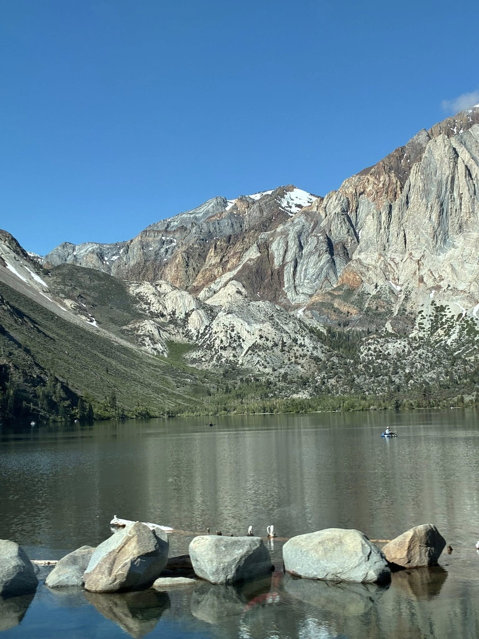 Mountain lake with shoreline rocks, calm water, and steep cliffs beneath a blue sky